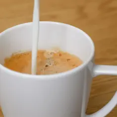 Biała filiżanka z kawą, do której wlewane jest mleko. Idealny napój na początek dnia, z uwzględnieniem kcal.
