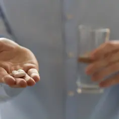 Dłoń trzyma dwie białe tabletki, w tym metioninę, obok szklanki wody.