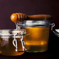 Dwa słoiki pełne złotego miodu, z drewnianej chochli spływa słodka kropla. Różne rodzaje miodów zachęcają do degustacji.
