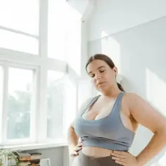 Kobieta zmagająca się z otyłością w sportowym stroju, z rękami na biodrach, stoi przed oknem.