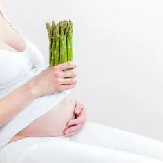 Kobieta w ciąży trzyma pęczek zielonych szparagów, symbol zdrowej diety.