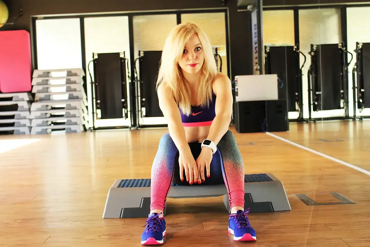 Blondynka w sportowym stroju siedzi na stepie, gotowa do wykonania 7 ćwiczeń na płaski brzuch.