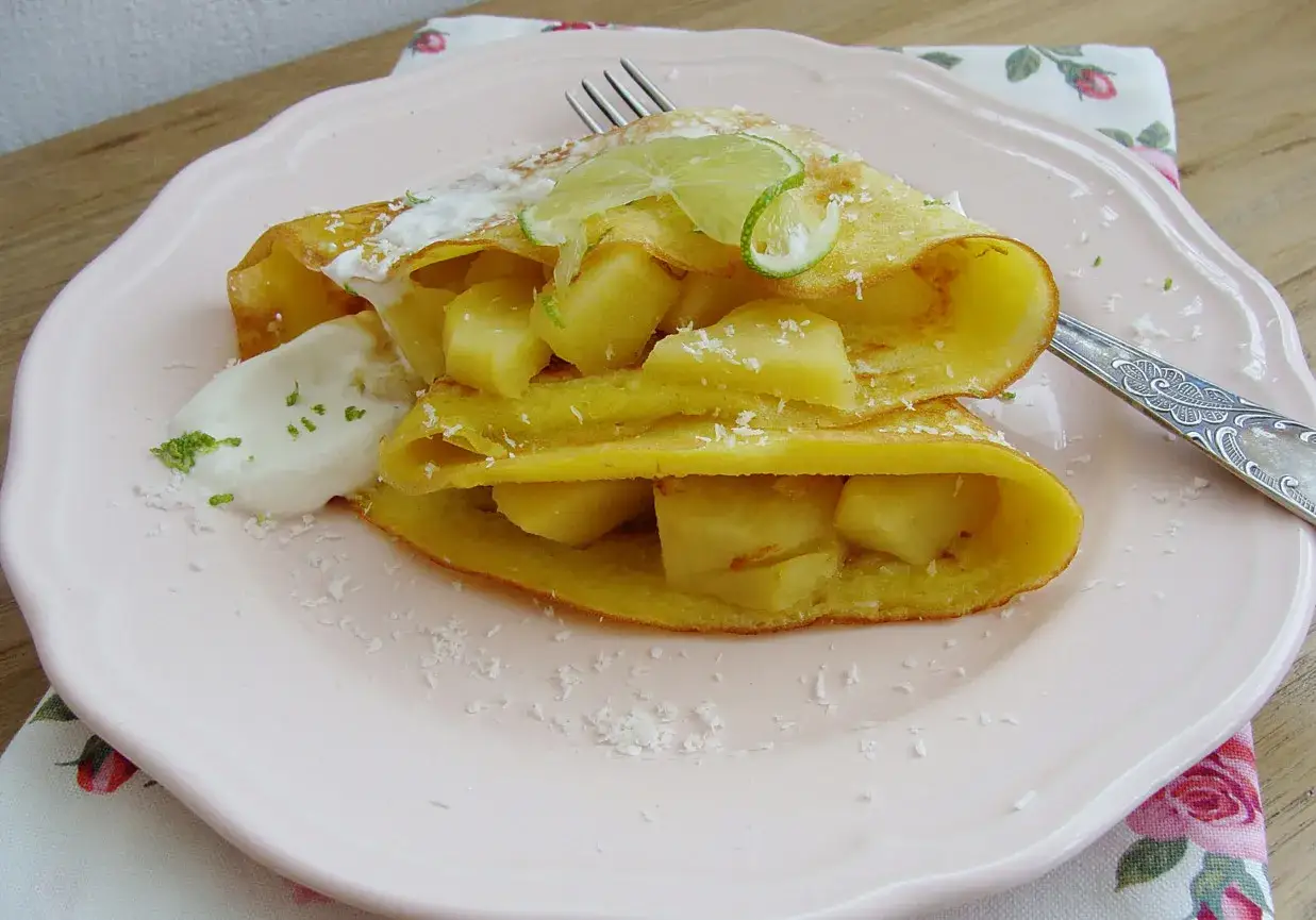 Naleśniki z mąki kokosowej z kawałkami ananasa, bitą śmietaną i plasterkami limonki. Posypane wiórkami kokosowymi.