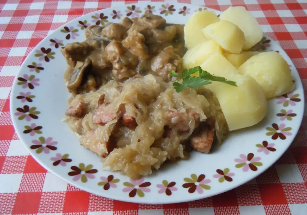 Na talerzu porcja kapusty kiszonej gotowanej z kiełbasą, obok ziemniaki i gulasz z grzybami.
