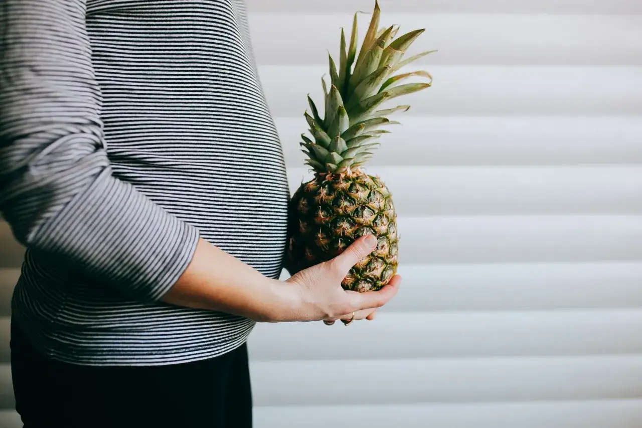 Brzuch kobiety w paski, trzymająca ananasa. To symboliczny ananas w ciąży, zapowiadający słodkie owoce.