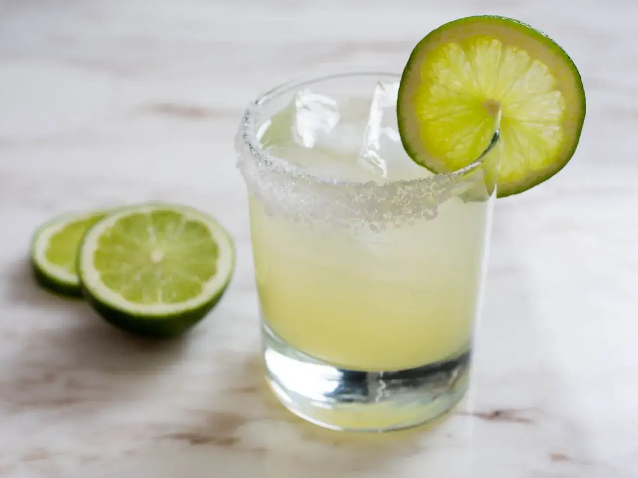 Orzeźwiająca margarita z lodem, solnym rantem i plasterkiem limonki. Obok leżą dwie połówki limonki.